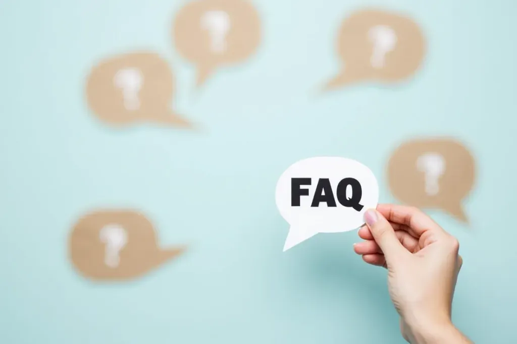 Hand hält eine Sprechblase mit der Aufschrift 'FAQ', im Hintergrund befinden sich mehrere Sprechblasen mit Fragezeichen.