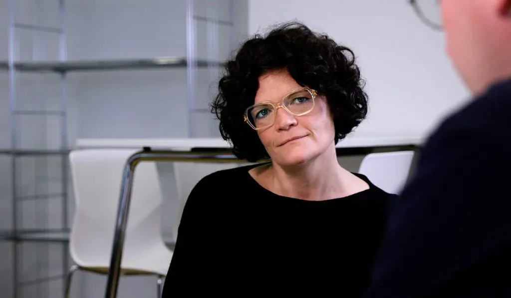 Eine Frau mit dunklen, lockigen Haaren und Brille sitzt in einem Raum und schaut nachdenklich zur Seite.