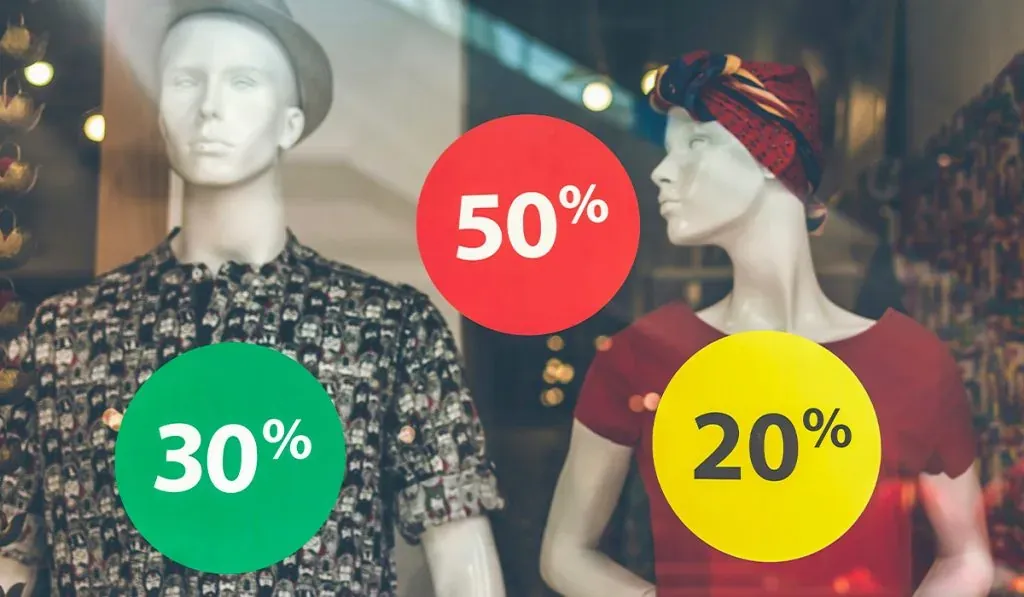 Schaufenster eines Bekleidungsgeschäfts mit zwei Schaufensterpuppen. Eine trägt ein gemustertes Hemd, die andere ein rotes Kleid und ein Kopftuch. Über den Puppen sind bunte Rabattaufkleber mit den Prozentzahlen 30%, 50%, und 20% in grün, rot und gelb angebracht.